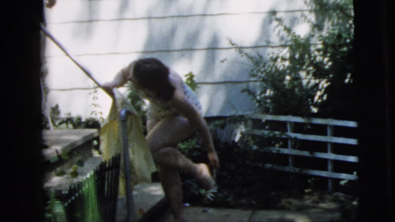 Young woman crouches near a garden gate with a towel and long-handled tool.