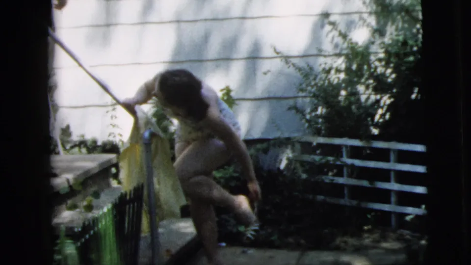 A young woman in a swimsuit crouches by a garden gate with a yellow towel and long-handled tool in the 1962 Camden backyard scene.