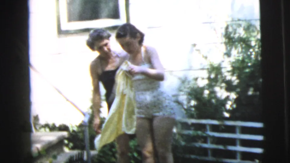 Two young women in swimsuits stand by the garden gate in the 1962 Camden backyard, with one holding a yellow towel.