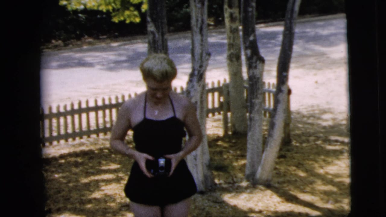 Woman in a black swimsuit stands outdoors holding a small camera near trees and a picket fence.