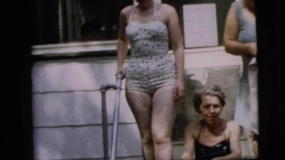 A standing woman in a patterned swimsuit and a seated woman on porch steps appear beside a visible crutch in 1962 home-movie footage.