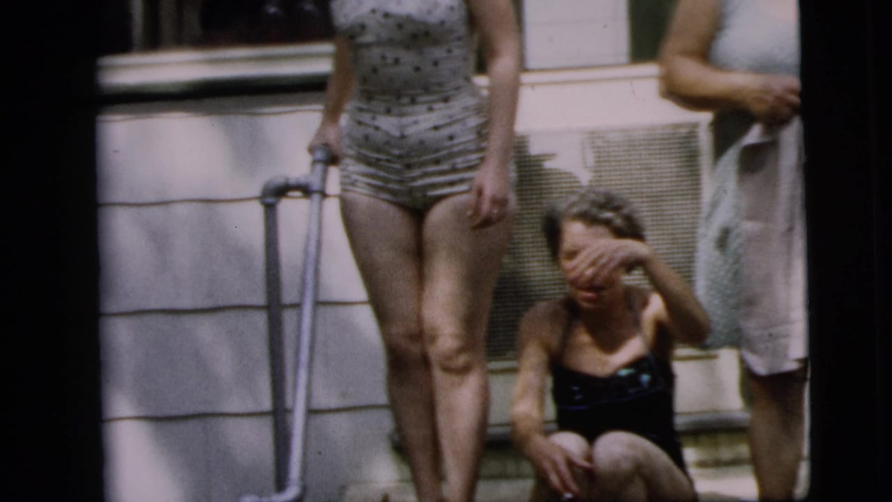 1962 Camden home-movie frame of adult women on a porch, with one woman in a polka-dot swimsuit using a crutch near a seated woman.