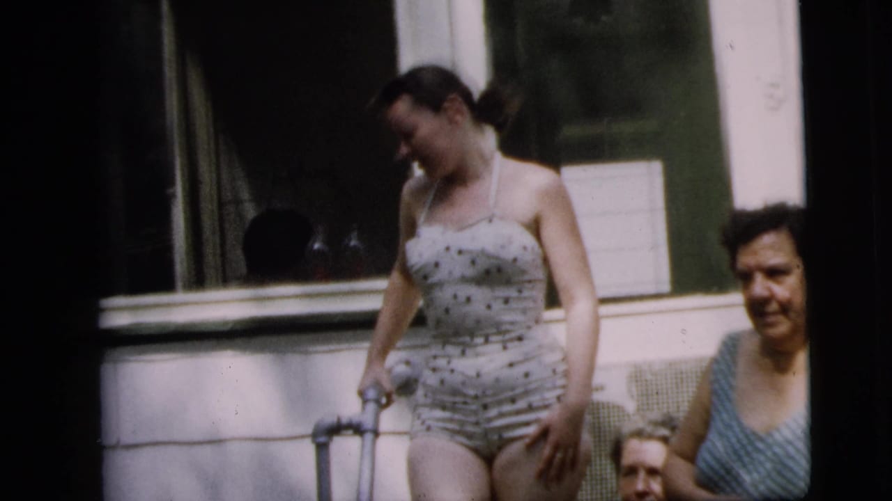 Adult woman in a polka-dot swimsuit stands with a crutch on a Camden porch, while other women sit or stand nearby in the 1962 footage.