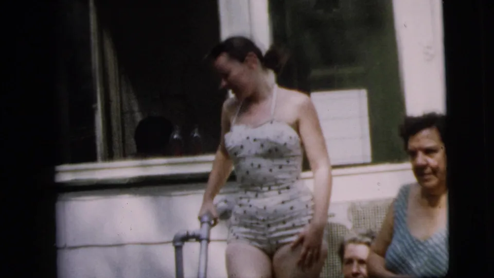Adult woman in a polka-dot swimsuit stands with a crutch on a Camden porch, while other women sit or stand nearby in the 1962 footage.
