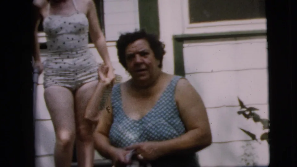 A seated older woman in a blue patterned swimsuit appears beside a partially cropped standing woman on a Camden porch in the 1962 home movie.