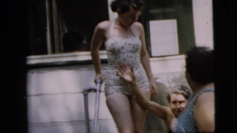 Adult women interact on a Camden porch, with the standing woman in a polka-dot swimsuit holding a crutch as others sit nearby in the 1962 clip.