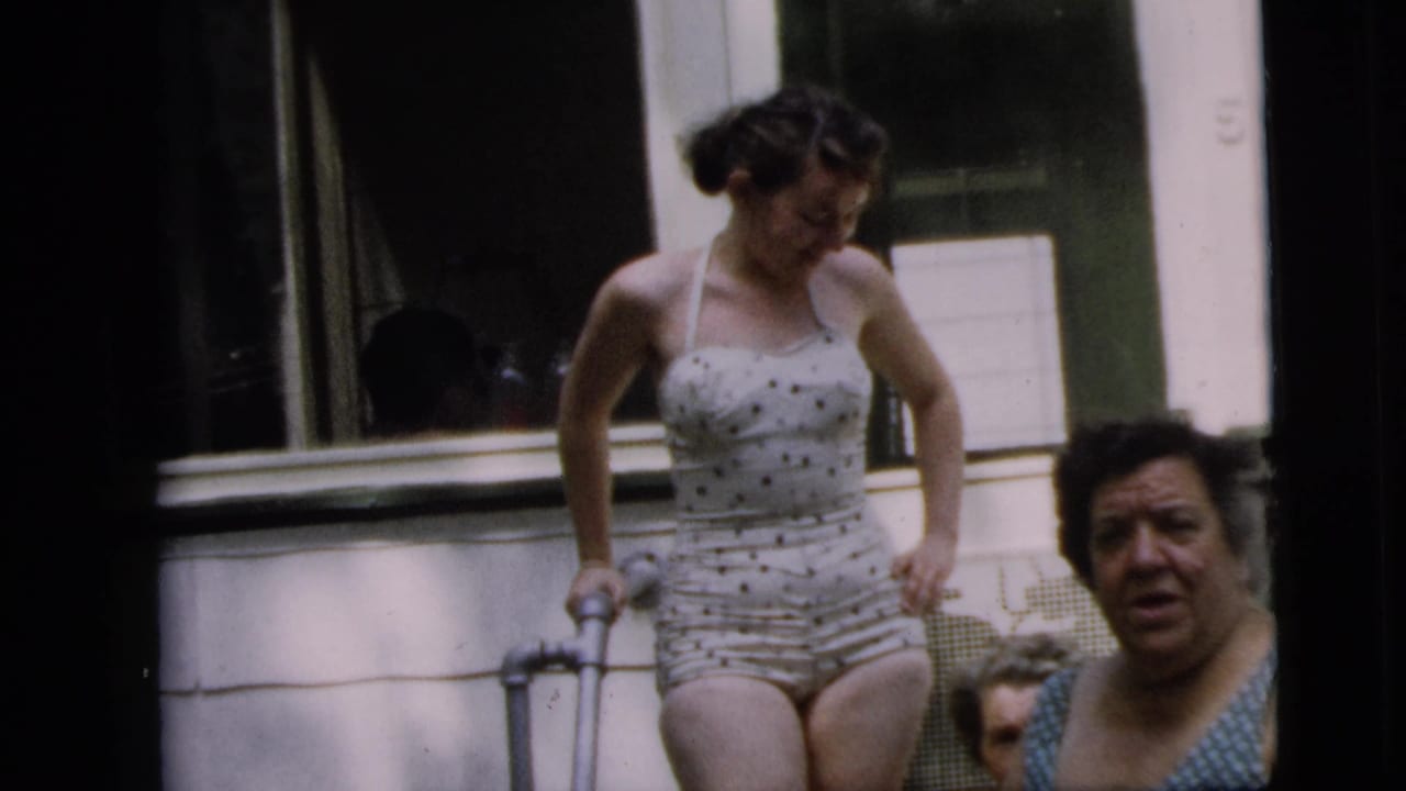 Woman in a polka-dot swimsuit uses a crutch on porch steps as another woman stands nearby.