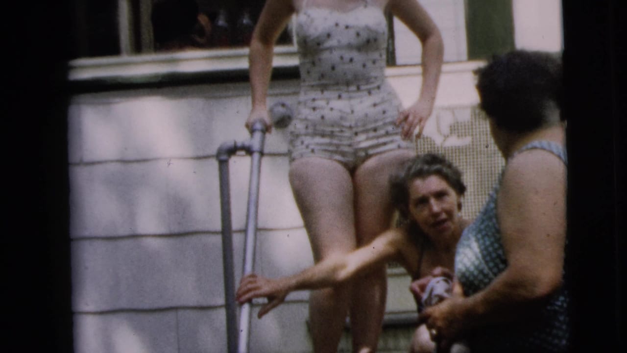 Woman with crutch stands on porch steps while two women in swimsuits gather near the doorway.