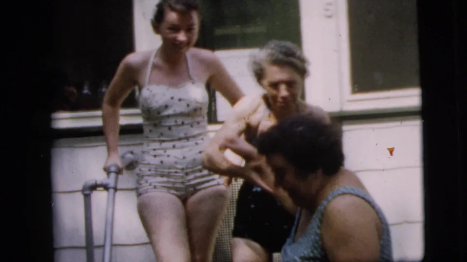 In the Camden porch scene, a woman in a polka-dot swimsuit stands behind an older woman in a dark swimsuit as another person moves through the foreground.