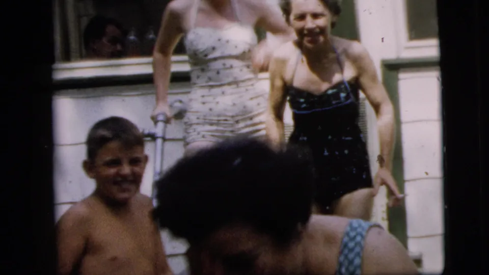 A young boy passes in front of Camden porch steps while women in one-piece swimsuits stand and move near the doorway and window.