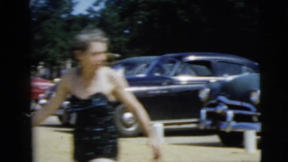 1962 Camden home-movie frame showing an older woman in a dark swimsuit walking in front of parked mid-century cars.