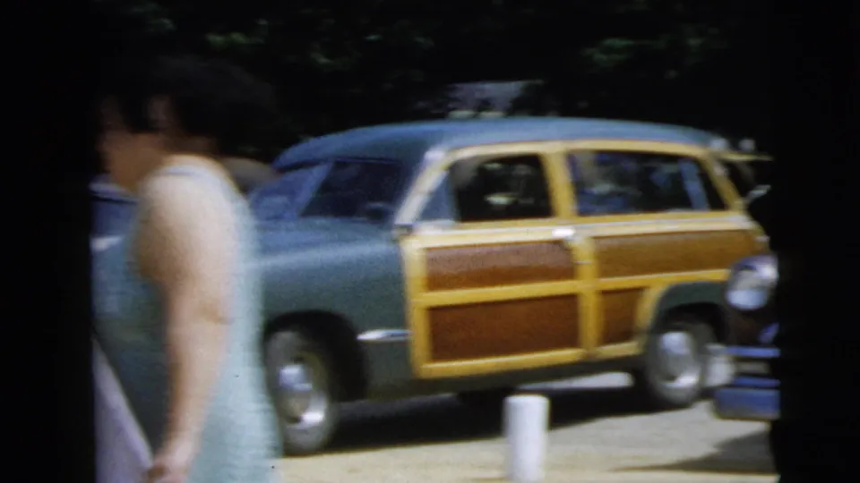 1962 Camden frame with a woman near a wood-paneled station wagon and other parked automobiles in bright outdoor light.