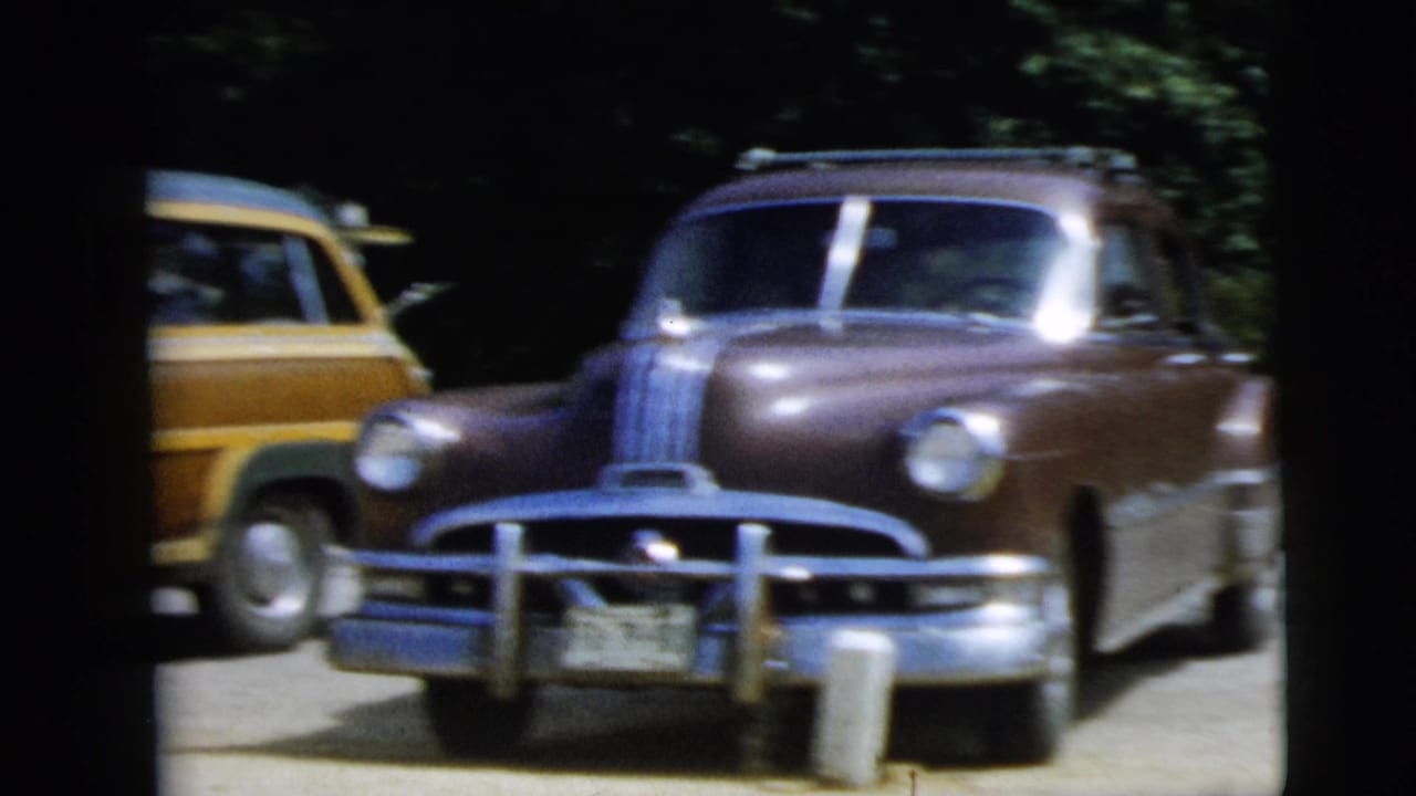 1962 Camden, New Jersey home-movie frame showing older American cars in a shaded parking area, including a wood-paneled station wagon.
