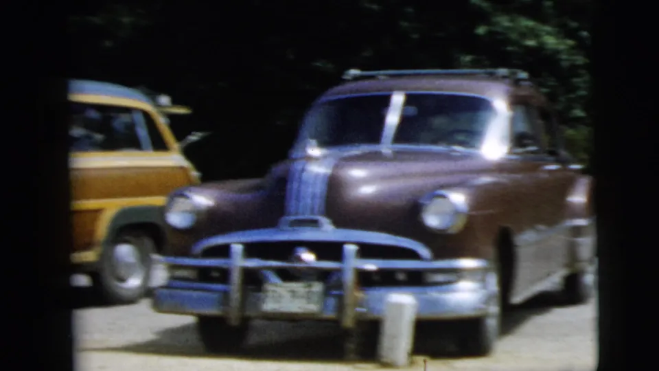 1962 Camden, New Jersey home-movie frame showing older American cars in a shaded parking area, including a wood-paneled station wagon.