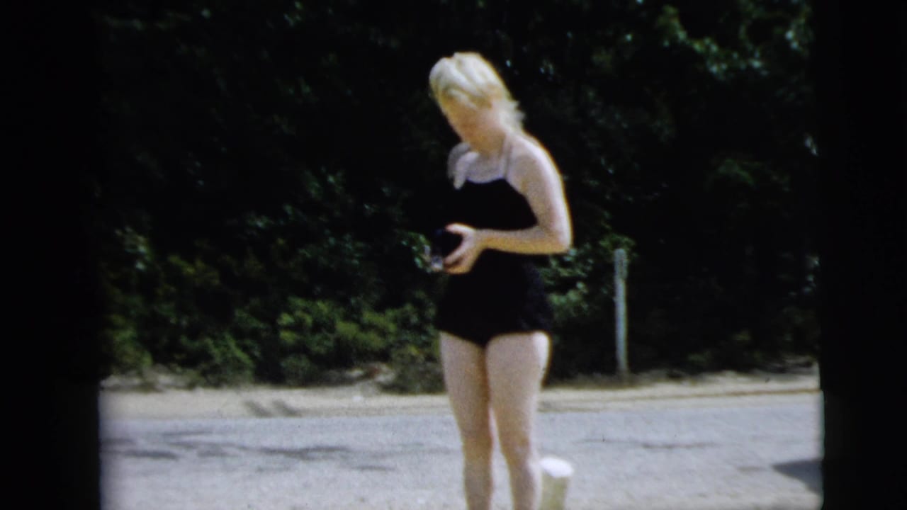 Woman in a black swimsuit stands near a sandy parking or lakeside area in the 1962 Camden home-movie footage, holding a camera.