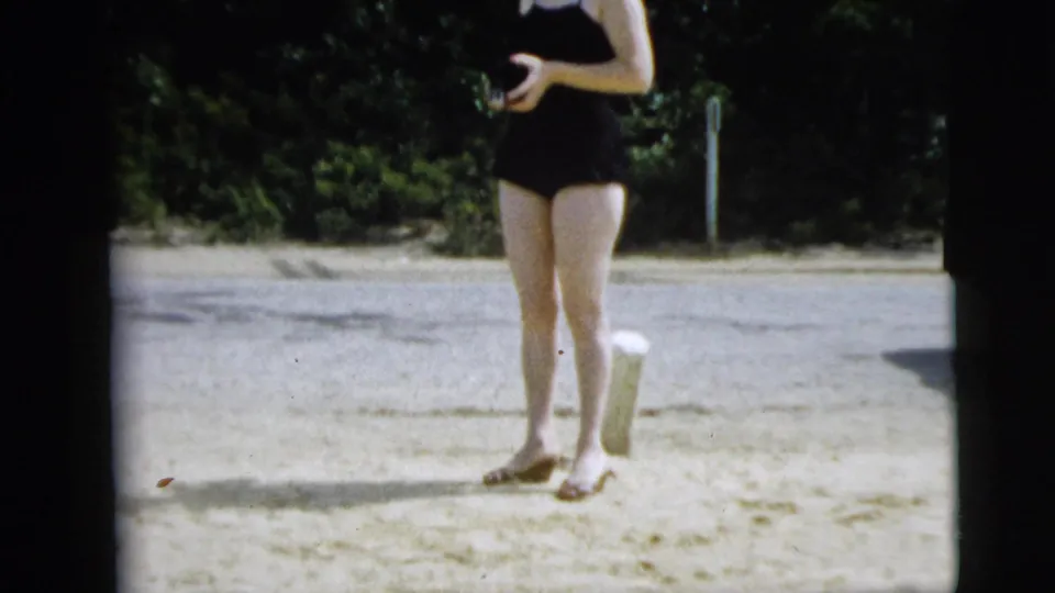 A woman in a black swimsuit is seen standing on sandy ground near trees in the Camden, New Jersey home-movie clip.