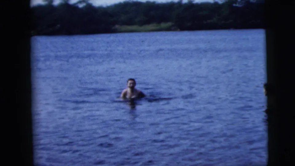 A swimmer is visible in calm lake water, with a wooded shoreline beyond, in the 1962 Camden-area home-movie sequence.