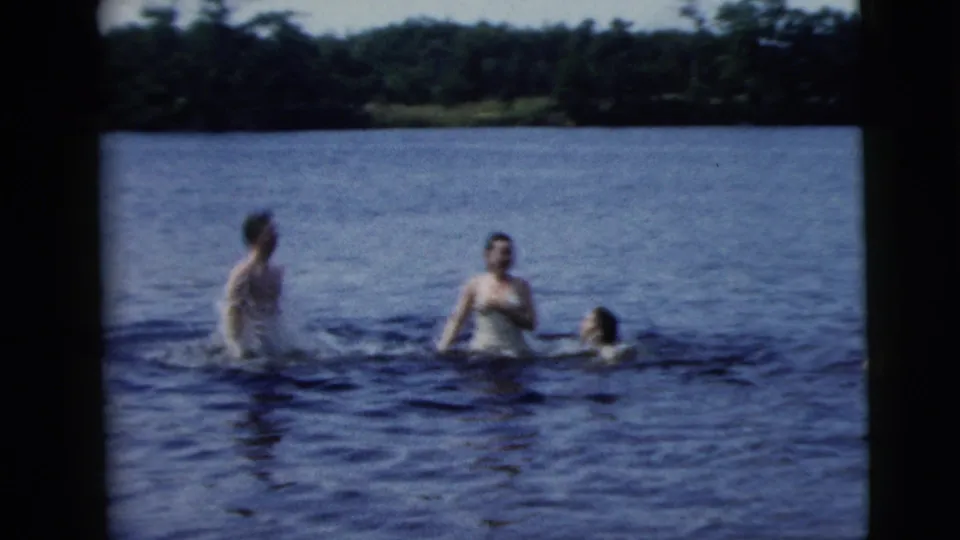 1962 home-movie frame from Camden, New Jersey metadata showing three adults in swimsuits standing and wading in lake water.