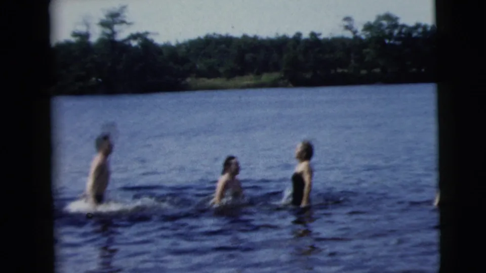 Three adults in swimsuits interact in a lake, with one person making a splash and a wooded shoreline in the background.