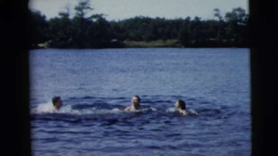 Home-movie frame of three adults swimming close to the waterline, with ripples and a distant tree-covered shore visible.