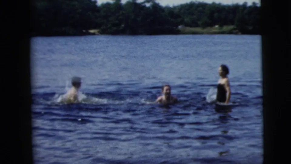 A 1962 lakeside home-movie frame shows two adults swimming and one adult standing in dark swimwear against open water and trees.