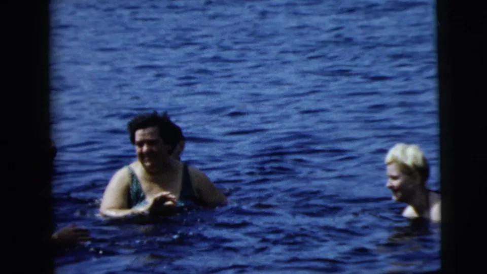 A woman in a swimsuit turns in the water while a blonde swimmer is visible at right, both framed by rippling blue open water.