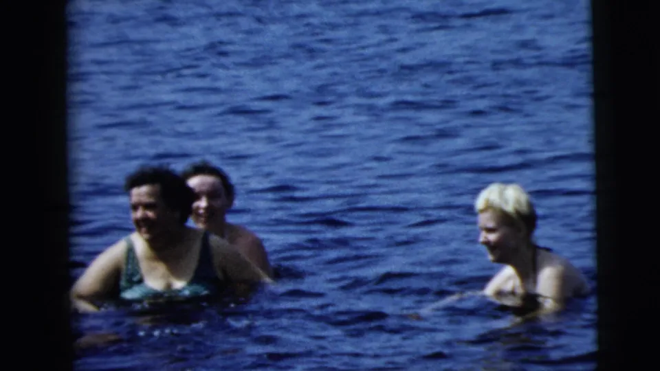 Three women are visible in the water, two smiling toward each other and the camera in a candid 1962 home-movie swimming scene.