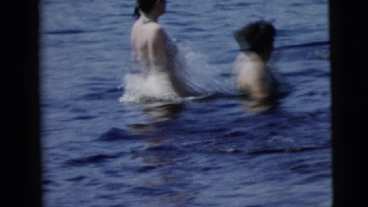 Two adult swimmers wade through open water in a soft 8mm frame, with one figure seen from behind and another at right.