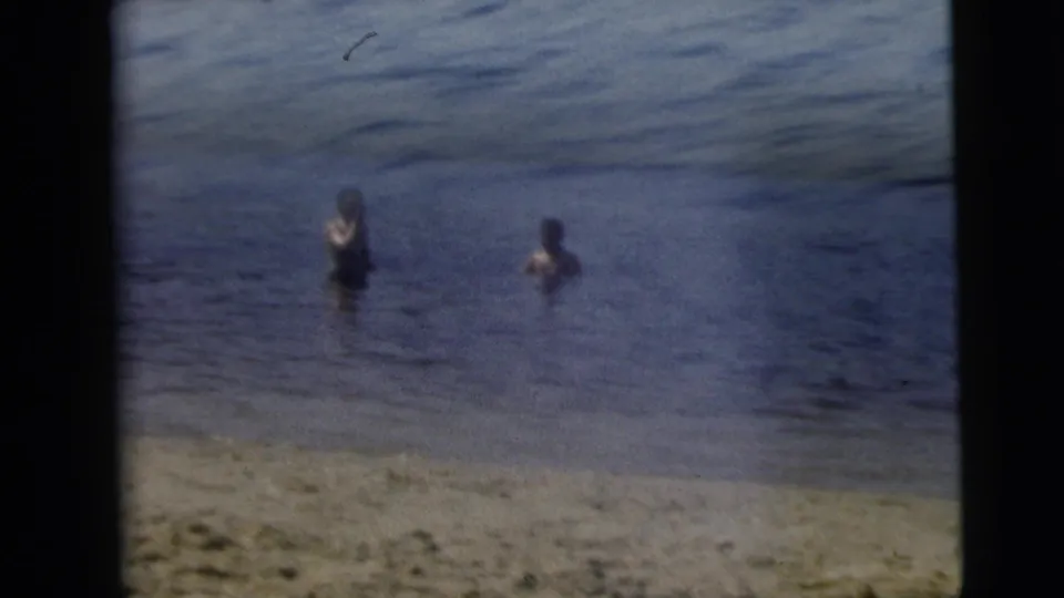 1962 Camden, New Jersey home-movie frame with two people in shallow lake water near a sandy shoreline, one raising an arm or object.