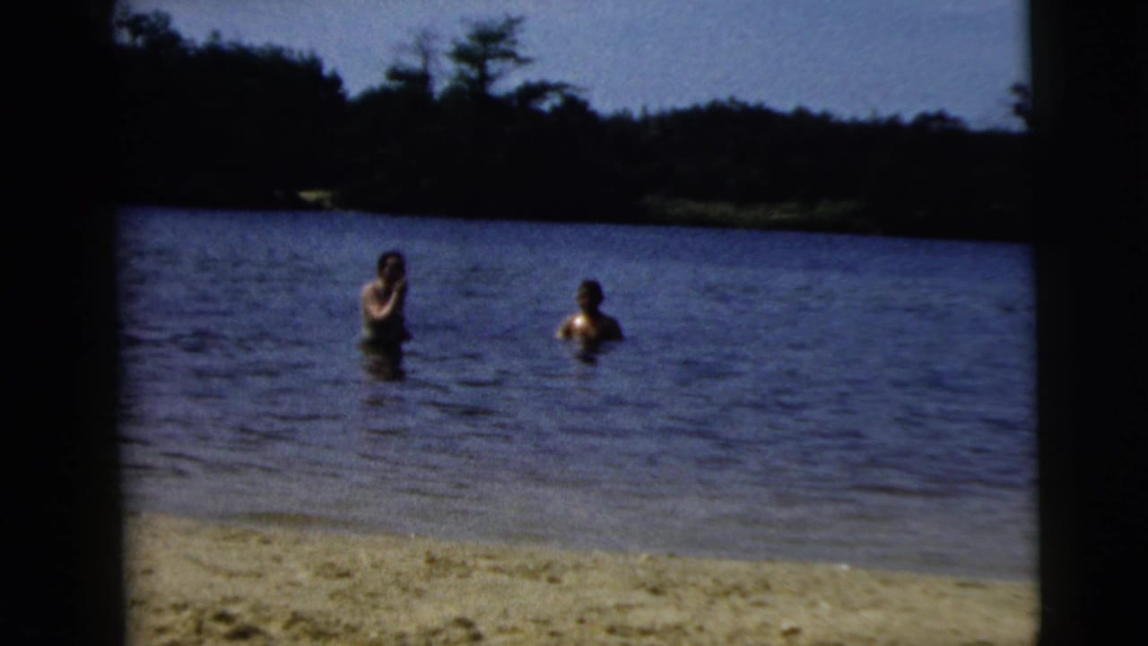 1962 8mm home-movie frame from Camden, New Jersey, showing two adults wading in a lake beside a sandy beach and wooded shoreline.