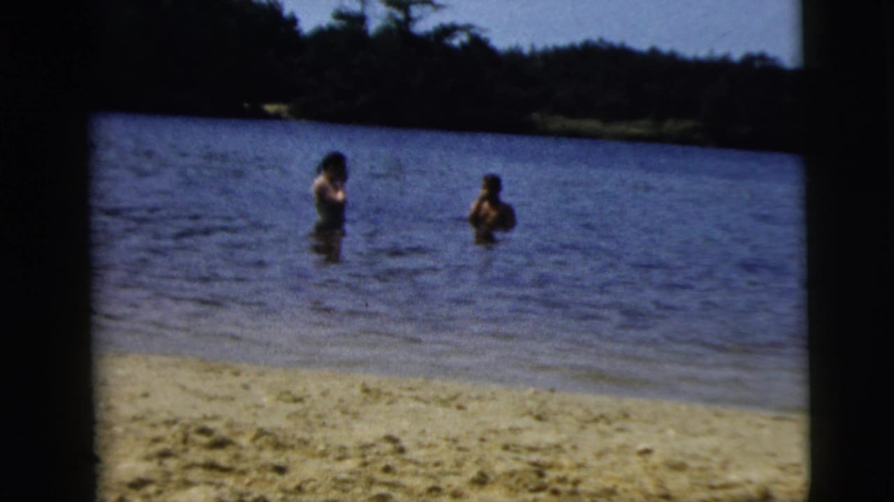 Camden, New Jersey 1962 home-movie frame with two adults standing in lake water beyond a sandy shoreline under a bright sky.