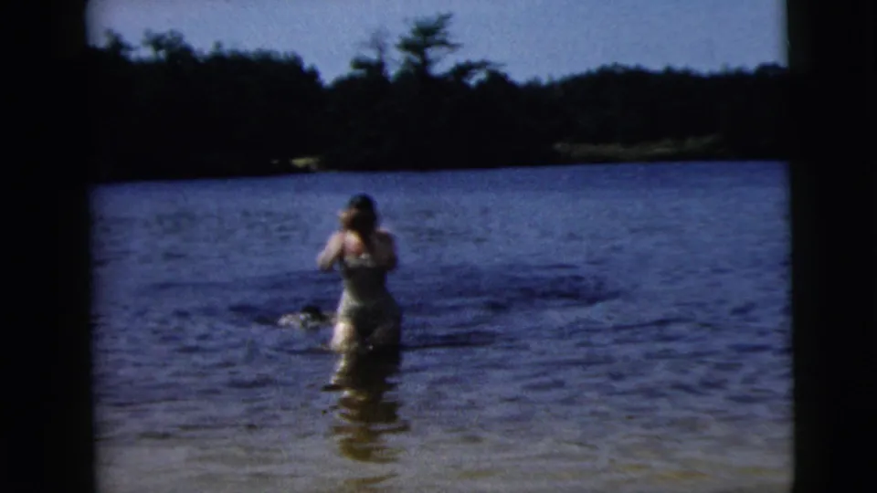 1962 Camden home-movie frame showing a woman in a one-piece swimsuit standing in shallow lake water near a wooded shore.