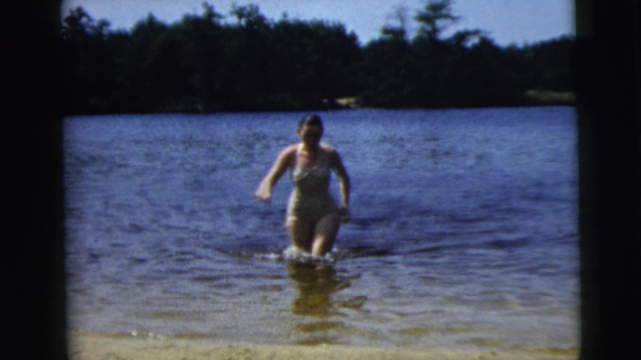 Woman in a one-piece swimsuit wading through shallow lake water toward the camera.