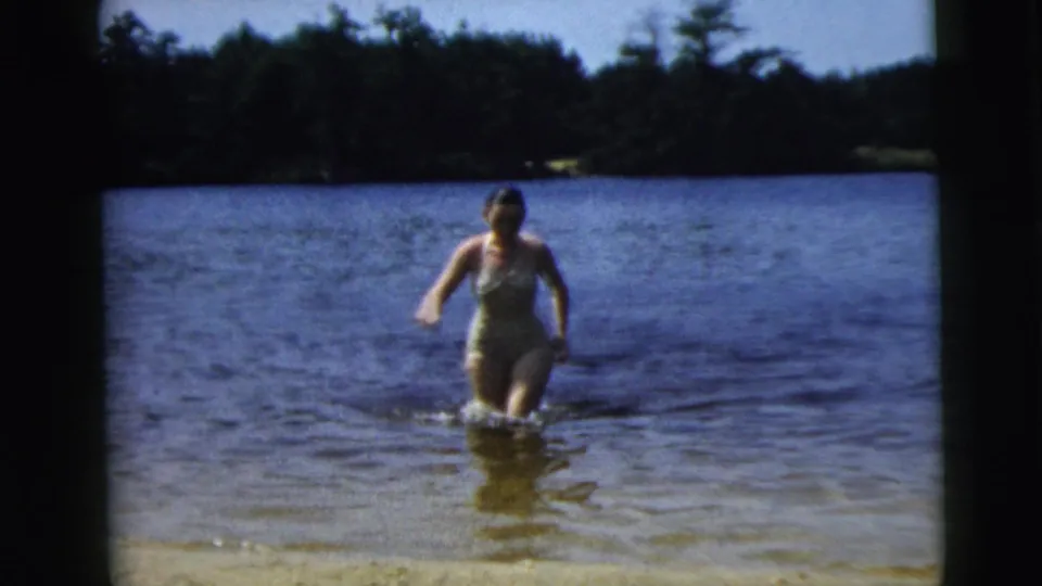 In supported 1962 Camden, New Jersey home-movie footage, a woman in a one-piece swimsuit wades from a lake toward shore.