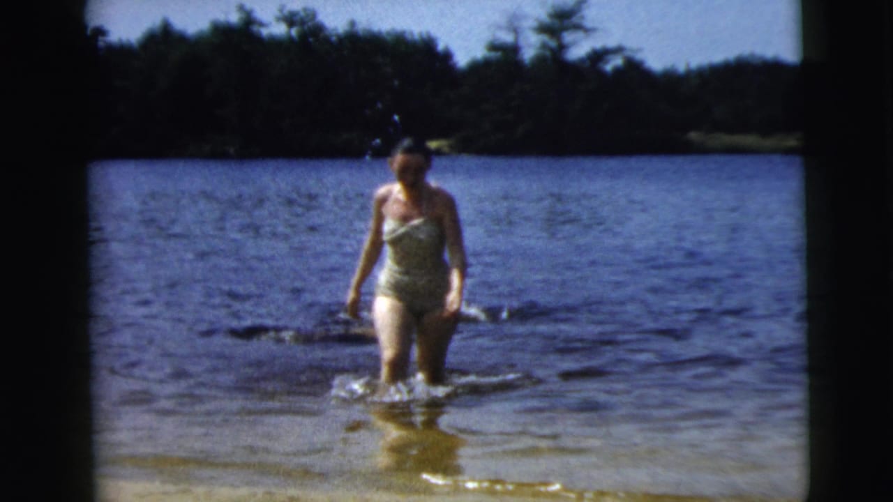 Woman in a swimsuit walking through knee-deep lake water near the shoreline.