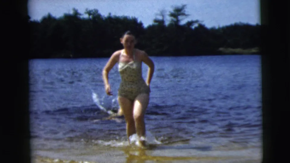 A woman in a patterned one-piece swimsuit steps through shallow lake water, with trees behind her in supported 1962 Camden footage.
