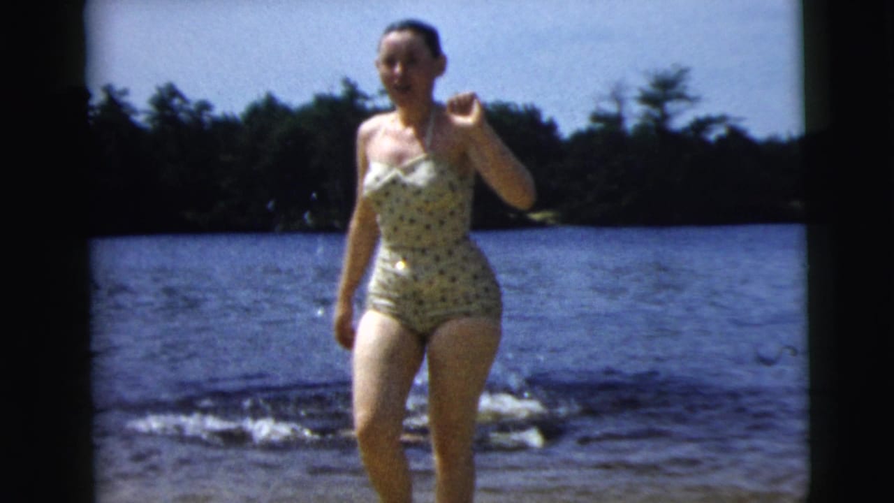 1962 Camden, New Jersey 8mm frame of a woman in a polka-dot one-piece swimsuit walking from a lake toward shore.