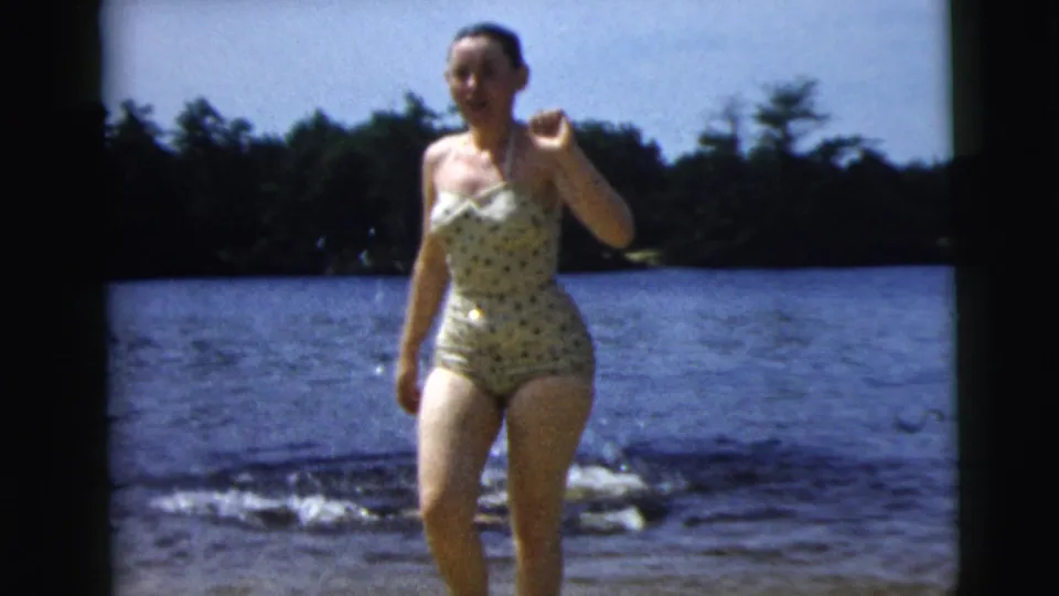 1962 Camden, New Jersey 8mm frame of a woman in a polka-dot one-piece swimsuit walking from a lake toward shore.