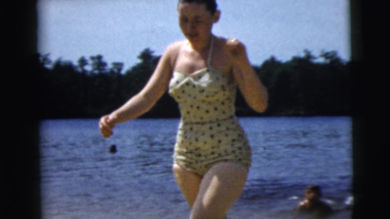 Woman in vintage polka-dot swimsuit walking near the lakeshore in supported 1962 Camden home-movie footage.
