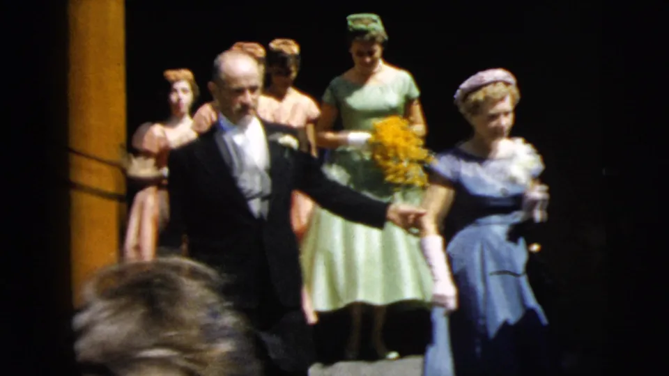 1962 Camden wedding home movie frame with a father in tuxedo escorting the bride in blue while pastel-dressed bridesmaids follow.