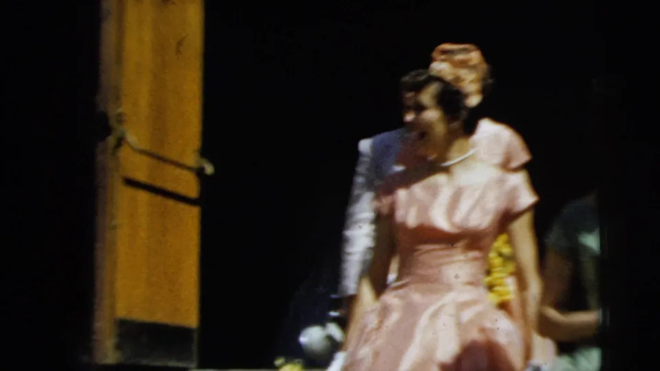 A woman in a pink formal dress appears near the doorway as the 1962 Camden group continues moving through the entrance area.
