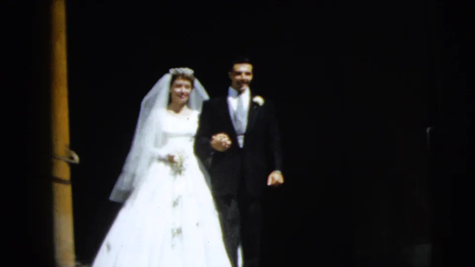 In 1962 Camden home-movie footage, a bride in a white gown and veil walks beside a groom in a dark suit near a dark doorway.