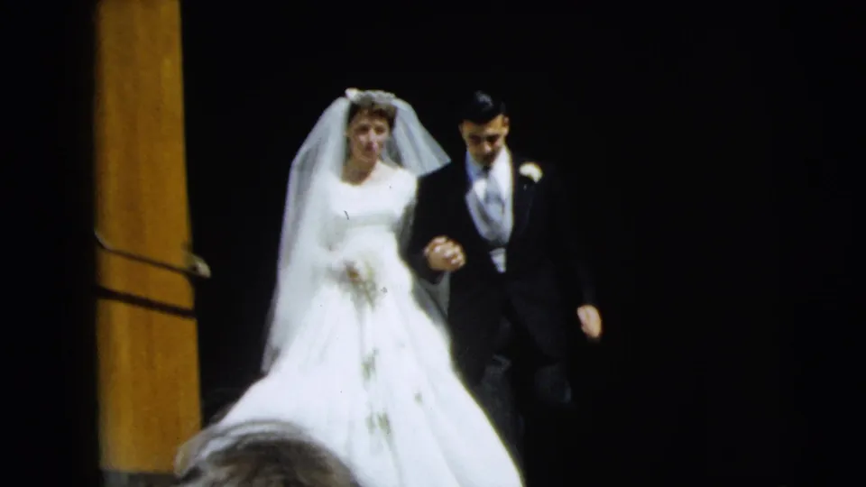 The couple walks down toward the camera, with the bride's full skirt visible and spectator heads partly entering the lower frame.
