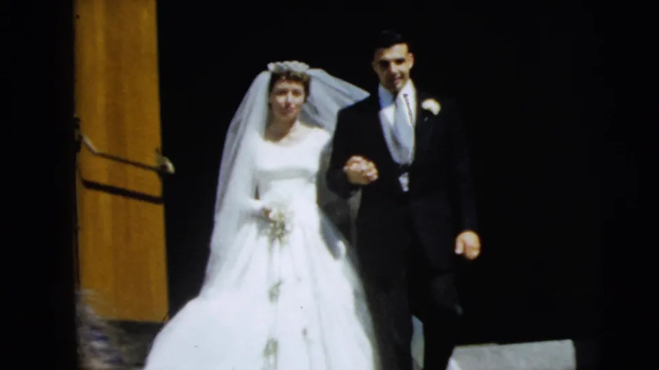 1962 Camden wedding home movie frame showing a smiling bride and groom descending steps, with a dark doorway behind them.