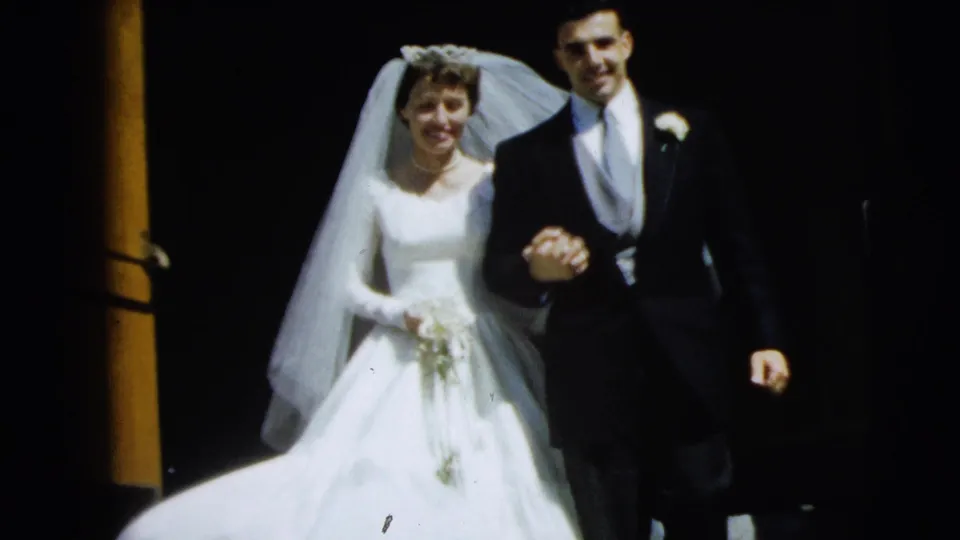 A close 8mm frame of the 1962 Camden newlyweds approaching the camera, with the bride's gown and veil prominent against the doorway.