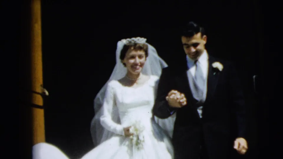 The bride smiles beside the groom near the end of the 1962 Camden wedding clip, with their hands joined in front of a dark entryway.