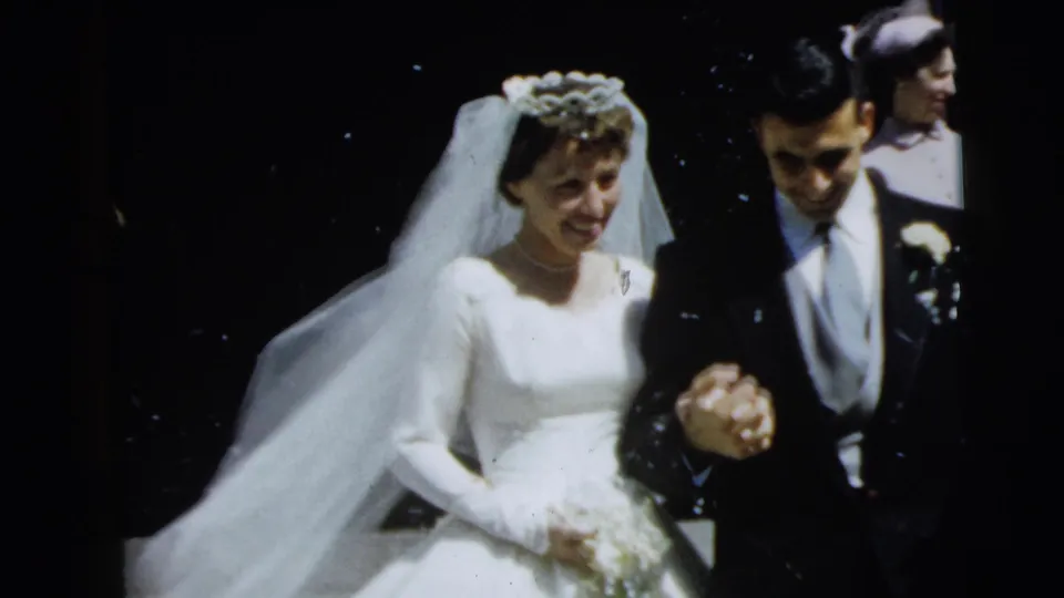 1962 Camden wedding scene with the bride smiling beside the groom as they walk past guests.