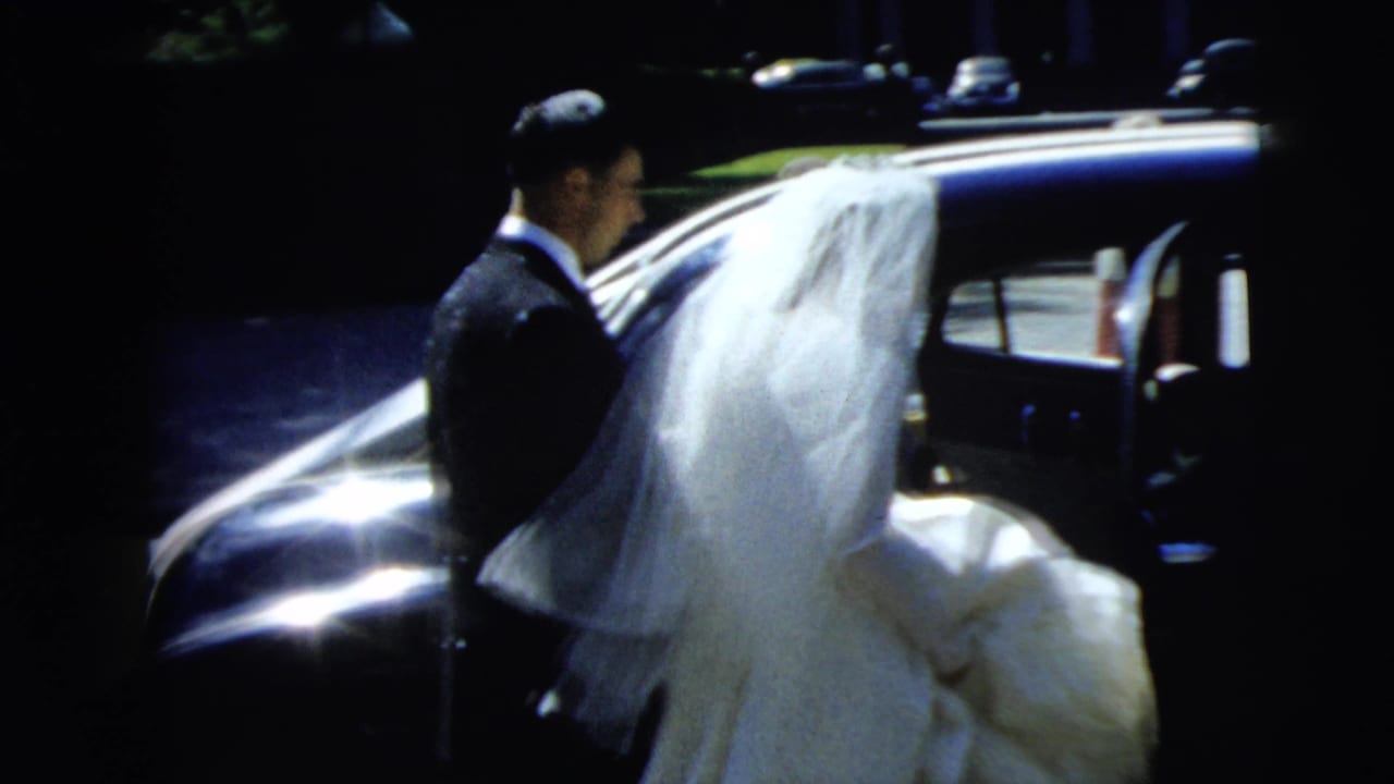 The groom in a dark suit stands by a classic sedan as the veiled bride approaches the car during the Camden wedding scene.