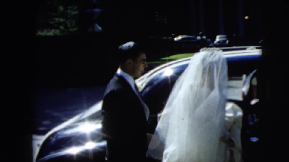 The groom stands near the bride's veil and the side of the sedan, with strong glare and shadow in the 8mm wedding footage.