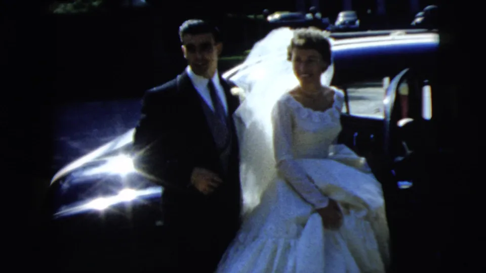 The newlywed couple, bride in white gown and groom in dark suit, stand beside the sedan in the 1962 Camden wedding footage.